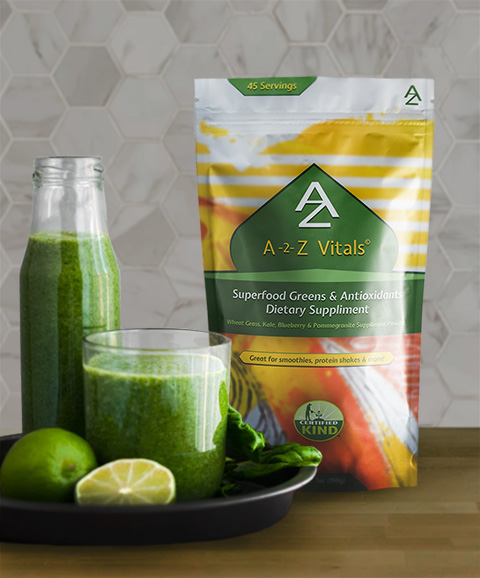 Yellow and green stand-up pouch labeled A-2-Z Vitals Superfood Greens & Antioxidants Dietary Supplement, 45 servings, next to a glass and bottle of green juice, limes, and greens on a black plate.
